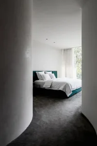 A minimalist bedroom with concrete walls and a bed with a green headboard and white bedding.