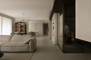 A minimalist living room with neutral-colored furniture and walls, featuring a lounge sofa, dining table, shelves, and a glas