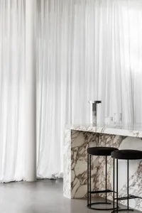 Bar stools placed at a marble counter in front of white curtains