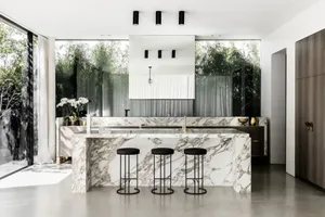 Modern kitchen featuring marble countertops and stools, with large windows showcasing greenery.
