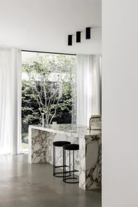 Modern kitchen with marble countertop and black stools, featuring large windows with white curtains.