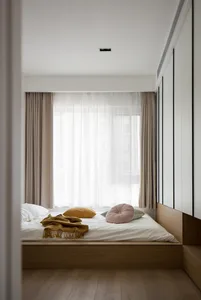 A minimalist bedroom featuring a low wooden bed with soft cushions and a blanket, under large windows with sheer curtains.