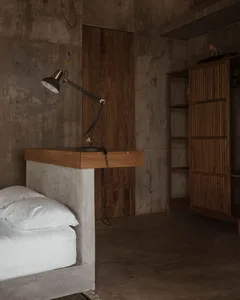 A bedroom with industrial design features a concrete bed frame and wooden desk with a table lamp.