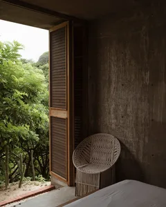 A modern bedroom featuring a concrete wall and a macrame chair, with a view of lush greenery outside through an open window.