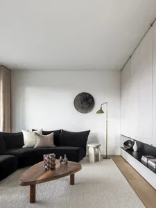A modern living room featuring a black L-shaped sofa with beige and white cushions, a wooden coffee table, and a concrete flo