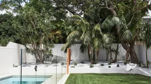 Modern poolside area with white stonework, glass fencing, and lush greenery including palm trees.