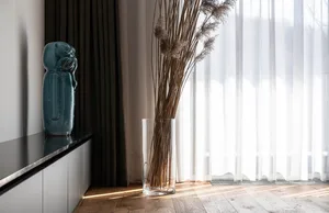 A tall cylindrical glass vase sitting on the floor between curtains and a window, containing dried grasses.