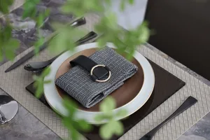 A modern table setting featuring a gray and white place mat, a black leather belt buckle napkin holder, and a gold ring detai