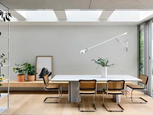 A minimalist dining room with a white table, cane chairs, and a white wall with skylights.