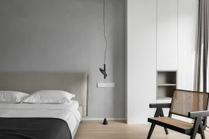 Modern minimalist bedroom featuring a bed with a gray fabric headboard, a hanging pendant light, and a woven cane chair.