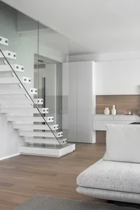 A minimalist apartment living room featuring a white staircase with glass railings, wooden flooring, and white furniture.
