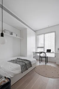 A minimalist white bedroom featuring a bed with a gray patterned blanket, a desk extension with a laptop, and a round rug.