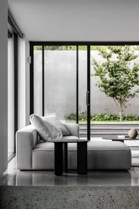 A minimalist living room with a gray modular sofa, black side table, and a view of a tree through large windows.