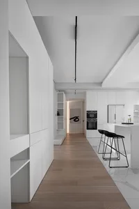 A modern, minimalist kitchen with white marble countertops, wooden flooring, and metal stools.