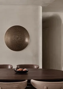 A minimalist dining room with a wooden table and chairs, featuring a round wall art piece with perforations and a bowl of app