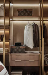 Modern walk-in closet with wooden shelves, hangers, and drawers, featuring a Louis Vuitton-style suitcase and hat.