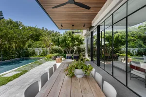 Modern patio area with a wooden dining table, white chairs, and a green plant, surrounded by a pool and lush greenery.