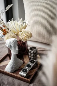 A decorative table display featuring a wooden tray, a ceramic bust sculpture, dried flowers, and metal candle holders on a be