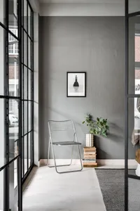 A minimalist living space featuring a metal chair, green plant, and book stack against a grey wall with a black frame window.