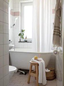 Minimalist bathroom with white tiled walls, stone flooring, and a freestanding bathtub under a window with a white shower cur
