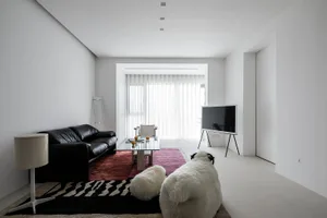 A minimalist living room with concrete walls and floor, featuring a black leather sofa, glass coffee table, and a television