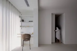 A minimalist residential interior featuring a white concrete wall, white floor, and a modern desk with shelves and a chair.