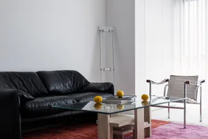 A modern minimalist living room with a black leather sofa, a glass coffee table, and a single glass chair next to a window wi