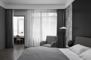 A modern minimalist bedroom with light gray walls, a bed covered in light gray bedding, a patterned gray armchair, and sheer 