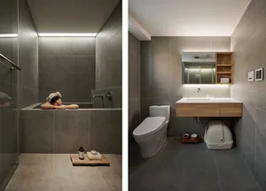 A modern bathroom featuring a gray tiled shower area with a person lying in a tub, a white toilet, and a vanity with a wooden