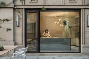 A modern kitchen with large glass walls, featuring a woman chopping vegetables on a counter.
