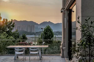 A minimalist balcony with wooden table, white chairs, glass railing, and concrete pillars overlooking mountains and cityscape
