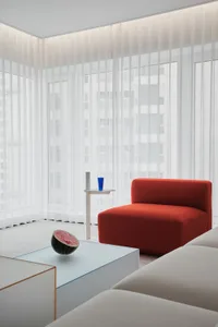A modern living room with an orange upholstered sofa, white curtains, and a glass table with a half-cut watermelon and blue c