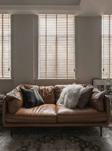 A modern leather sofa adorned with throw pillows and placed in front of a window with wood blinds.