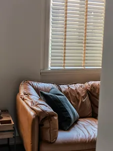 A modern leather sofa with pillows sits by a window with blinds.