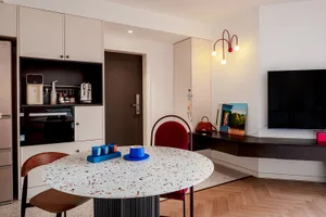 A minimalist kitchen featuring a round, speckled terrazzo table, red and brown chairs, and modern lighting fixtures.