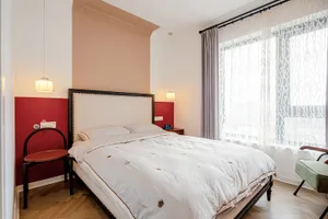 A minimalist bedroom with a white bed, pendant lights, red accent wall, and large window with sheer curtains.