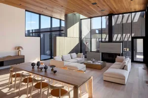 A modern living room featuring wooden furniture and flooring, with large windows allowing natural light to fill the space.