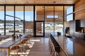 Modern interior featuring wood and black steel dining and kitchen setup with large windows overlooking a snowy landscape.