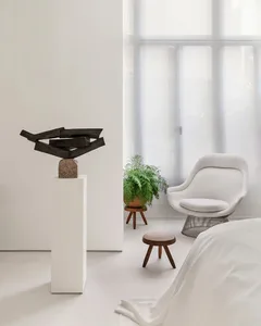 A minimalist bedroom featuring a white bed with a modern sculpture on a pedestal and a white chair with a green plant.