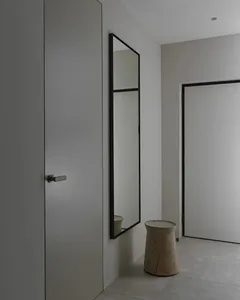 A minimalist entryway featuring a wooden stool, white walls, and a large mirror with a black frame.