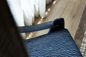 A close-up of a blue textured cloth leather chair with a wooden floor and a white curtain in the background.