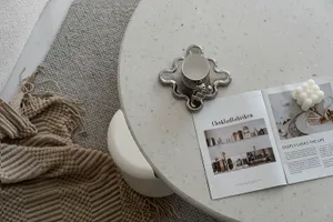 A round white terrazzo coffee table with a white ceramic cup, a metallic candle holder, and an open magazine on the table.