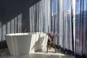 A minimalist bathroom featuring a white terrazzo tub and cotton-blend curtains, illuminated by natural light.