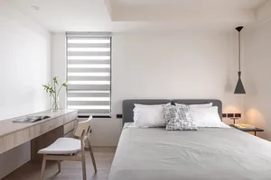 A minimalist bedroom with a fabric bed, wooden desk, and roller blinds.