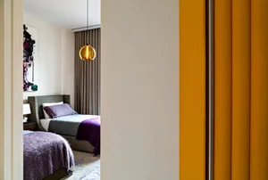A modern bedroom with a purple blanket on the bed, featuring a wicker bed frame, a glass pendant light, and grey and purple b