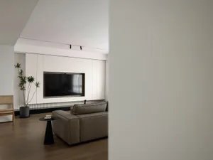 A modern minimalist living room featuring a grey sectional sofa, a black side table, a flat-screen TV embedded in a white pan