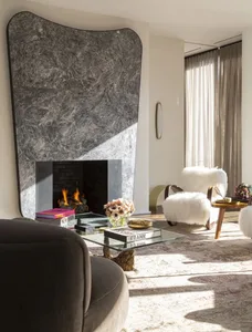 A cozy living room with a gray marble fireplace and a fuzzy white armchair.