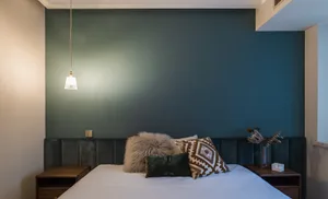 A minimalist bedroom featuring a bed with a velvet headboard, decorated with various pillows and a fur blanket, illuminated b