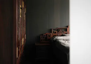 A minimalist bedroom with a wooden headboard, neatly made bed, and dark wooden furniture against a dark grey wall.