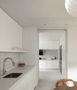A minimalist kitchen with white cabinets, a stainless steel sink, and a light fixture hanging above the dining area.